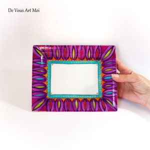 May include: A rectangular ceramic plate featuring a white center bordered by a bright turquoise frame with gold dots. The outer edge is decorated with a repeating pattern of purple, lime green, and orange petal shapes. The text De Vous Art Moi is visible.
