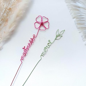 Pode incluir: Três peças de arte em arame sobre um fundo branco. Uma é uma flor rosa, outra diz "maîtresse" em arame rosa, e a terceira diz "merci" em arame verde com um desenho de folha. As peças são delicadas e feitas à mão.