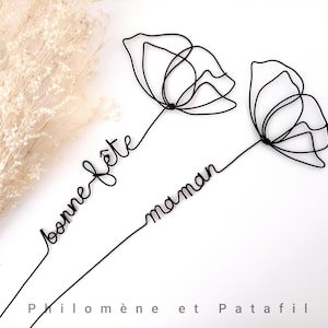 Pode incluir: Arte floral em arame preto com as palavras "bonne fête maman". O design delicado apresenta duas flores estilizadas com caules finos e o texto em escrita cursiva. O nome do artista, "Philomène et Patafil", está na parte inferior.
