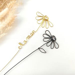 Pode incluir: Duas decorações de flores de margarida de arame. Uma é dourada com a palavra "Maman" em arame dourado. A outra é preta. Ambas têm um caule e pétalas. O fundo é branco.