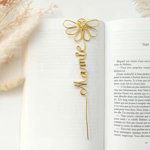 Könnte beinhalten: Ein goldfarbener Draht-Lesezeichen in Form einer Gänseblume mit dem Wort "Mamie" in Schreibschrift. Das Lesezeichen befindet sich in einem offenen Buch. Getrocknete Blumen sind im Hintergrund. Das Lesezeichen ist ein dekorativer Artikel.
