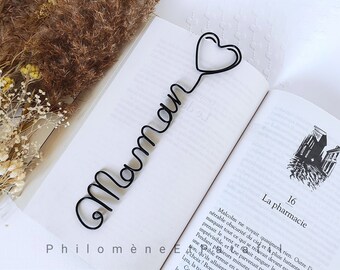 May include: Black wire bookmark in the shape of a heart with the word "Maman" in cursive script. The bookmark is placed in an open book. The book's pages are white, and the text is in French.