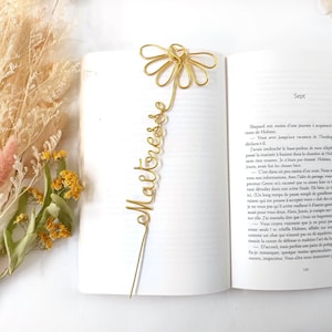 Puede incluir: Un marcapáginas de alambre de color dorado con forma de flor en la parte superior y la palabra "Maîtresse" escrita en cursiva a lo largo del tallo, colocado sobre las páginas abiertas de un libro con texto impreso en francés.