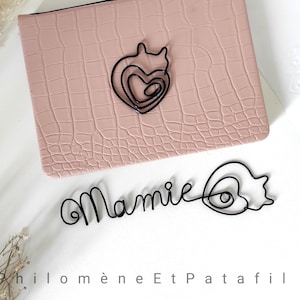 Op de afbeelding: Een roze, getextureerde portemonnee met een zwart draad hart ontwerp en het woord "Mamie". Het ontwerp staat op een wit oppervlak met gedroogde bloemen en de tekst "Philomène Et Patafil".