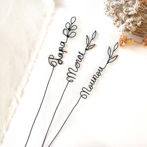 Peut inclure: Trois tiges en fil de fer noir avec des inscriptions en écriture cursive. Les tiges portent les mots Papa, Merci et Nounou, surmontés de motifs de feuilles en fil métallique. Le fil est fin et sombre, formant des boucles continues.