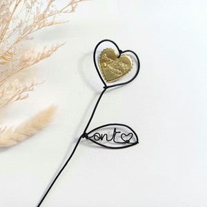 Puede incluir: Decoración en forma de corazón de alambre negro con un corazón dorado en el centro que dice "Bonne Fête Maman". Una forma de hoja de alambre negro con la palabra "ont" y un corazón debajo.