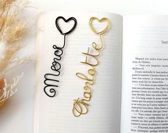 Op de afbeelding: Twee draadboekenleggers op een open boek. De ene boekenlegger is zwart en bevat het woord Merci in cursief schrift met een hartje erboven. De andere boekenlegger is goudkleurig en bevat het woord Charlotte in cursief schrift met een hartje.