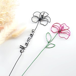Puede incluir: Dos flores de alambre, una negra y otra rosa, con tallos y hojas verdes. La flor negra tiene la palabra "Maman" en cursiva. Las flores están sobre un fondo blanco con flores secas en la esquina superior izquierda.