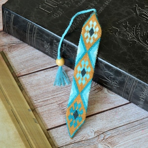 May include: A handmade bookmark with a geometric pattern in shades of turquoise, orange, and black. The bookmark has a tassel and a wooden bead. The bookmark is resting on a book with a dark cover.