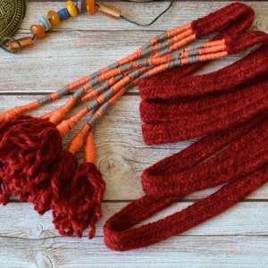 May include: A set of red yarn tassels with orange and gray accents. The tassels are attached to a long, red knitted cord.