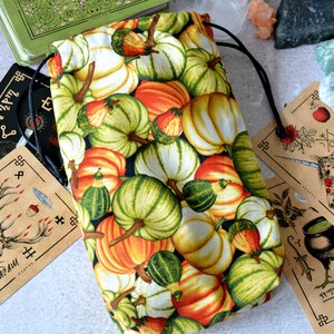 Op de afbeelding: Een trekkoordtas gemaakt van stof met een patroon van oranje, groene en witte pompoenen. De tas ligt op een tafel met andere items, waaronder kaarten met illustraties en kristallen.