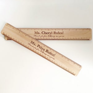 May include: Two wooden rulers with engraved messages. The top ruler says "Ms. Cheryl Rules! Thank you for helping me grow." The bottom ruler says "Ms. Priya Rules! Thank you for helping me grow."