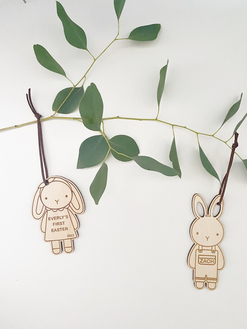 Puede incluir: Dos adornos de conejo de madera con colgadores de cuerda marr&oacute;n. El conejo de la izquierda es una coneja con un vestido y el texto "EVERLY'S FIRST EASTER 2022". El conejo de la derecha es un conejo con un mono y el texto "ZACH".