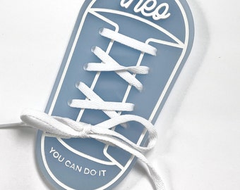 Puede incluir: Una herramienta de aprendizaje en forma de zapato azul claro con cordones blancos y el texto "You Can Do It" y el nombre "theo" en la parte superior.