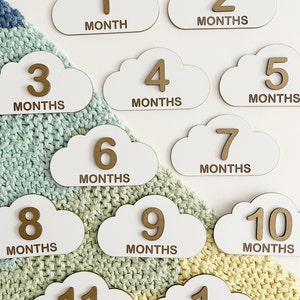 Pode incluir: Cartas de marcos de desenvolvimento para bebês de madeira em forma de nuvem, com números dourados e as palavras "mês" ou "ano" em cada cartão. Os cartões estão dispostos em um padrão em um cobertor de malha verde e amarelo.