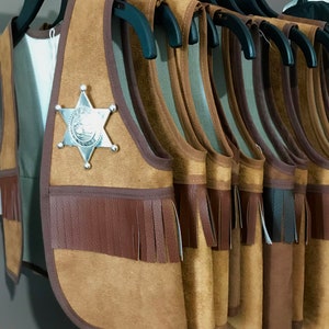 May include: A row of brown suede vests with fringe trim and a silver sheriff's star badge on the front vest. The vests are hanging on black hangers.