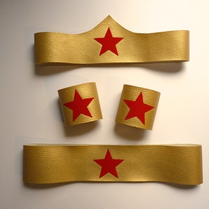 Pode incluir: Um conjunto de três acessórios de fantasia de Mulher Maravilha de cor dourada. O conjunto inclui uma faixa de cabeça, um par de punhos e um cinto. Cada peça apresenta uma estrela vermelha.