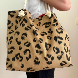 Puede incluir: Bolso tote grande con estampado de leopardo beige y asas de cuerda. El bolso presenta manchas negras sobre un fondo beige. Las asas son de cuerda blanca con borlas decorativas en la parte superior. El interior es rojo.