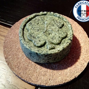 Peut inclure: Une barre de savon ronde de couleur verte avec un motif de trèfle à quatre feuilles. Le savon est posé sur un dessous de verre en liège brun. Le savon est étiqueté "Fabrication Française Fait Main".