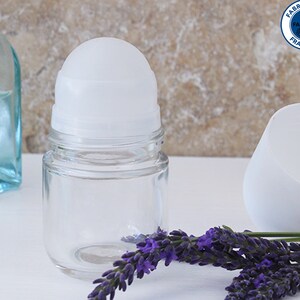 Peut inclure: Un flacon transparent en verre à bille avec un bouchon en plastique blanc et un capuchon en plastique blanc. Le flacon est posé sur une surface blanche avec une brindille de fleurs de lavande.