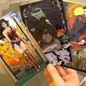 Könnte beinhalten: Drei Comic-Kunstdrucke mit den DC Comics-Superhelden Wonder Woman, Batman und Superman. Der Wonder Woman-Druck hat einen goldenen und orangenen Hintergrund. Der Batman-Druck hat einen dunklen Hintergrund. Der Superman-Druck hat einen blauen und goldenen Hintergrund.