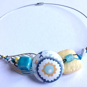 Puede incluir: Un collar hecho a mano con un alambre plateado y elementos decorativos. El collar incluye un botón grande estampado, cuentas turquesas, una hoja dorada y una cuenta amarilla clara en forma de corazón. El diseño incorpora acentos azules, amarillos y blancos.