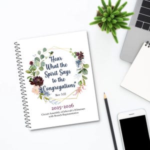 Op de afbeelding: Een witte spiraalgebonden planner met de tekst "Hear What the Spirit Says to the Congregations" en "2025-2026". De omslag heeft bloemen en geometrische ontwerpen. Een potlood, smartphone en kleine plant zijn ook zichtbaar.