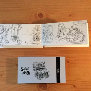 May include: A sketchbook with black and white illustrations of people and cityscapes. The cover features the word "Seoul" in English and Korean, along with a drawing of a building. The sketchbook is open, revealing detailed drawings.