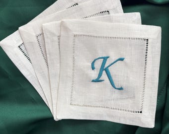 May include: Four white linen cocktail napkins with a teal embroidered letter "K" in the center. Each napkin has a decorative stitched border. The napkins are arranged on a dark green satin surface.