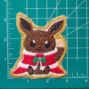 May include: A brown and tan cartoon character wearing a red and white Santa suit with a green bow tie. The character is embroidered on a gold fabric patch.