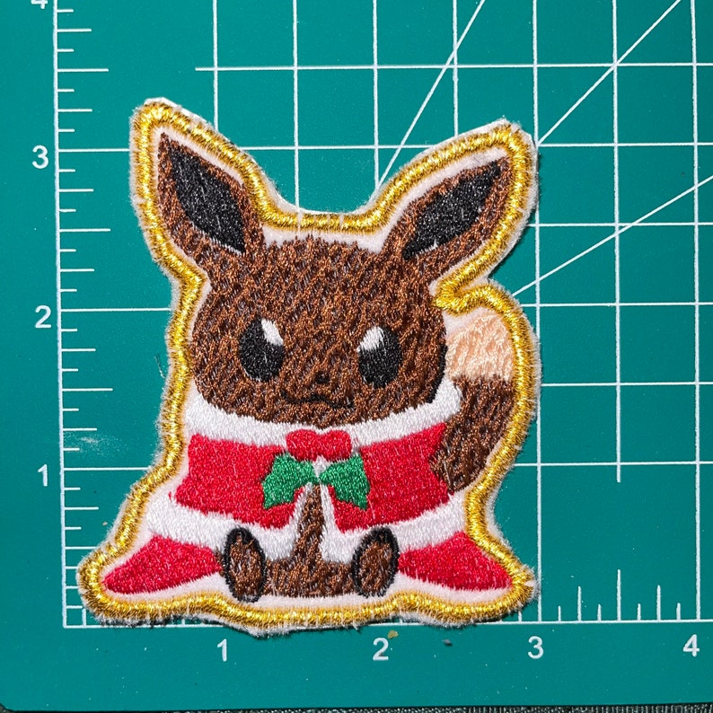 May include: A brown and tan cartoon character wearing a red and white Santa suit with a green bow tie. The character is embroidered on a gold fabric patch.