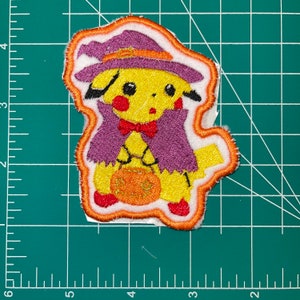 May include: An embroidered iron-on patch featuring a yellow Pikachu dressed as a witch holding a pumpkin. The patch has a purple cape, orange hat, and a white background.