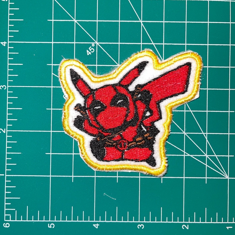May include: Embroidered patch featuring a mashup of Deadpool and Pikachu. The design shows Deadpool's mask and costume in red, with Pikachu's ears and tail. The patch has a white background with a gold border.