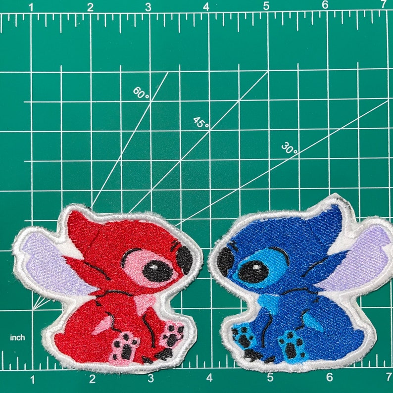 May include: Two embroidered patches of Stitch, a character from the Disney movie Lilo & Stitch. One patch is red and the other is blue. Both patches have white outlines and are sitting on a green grid.