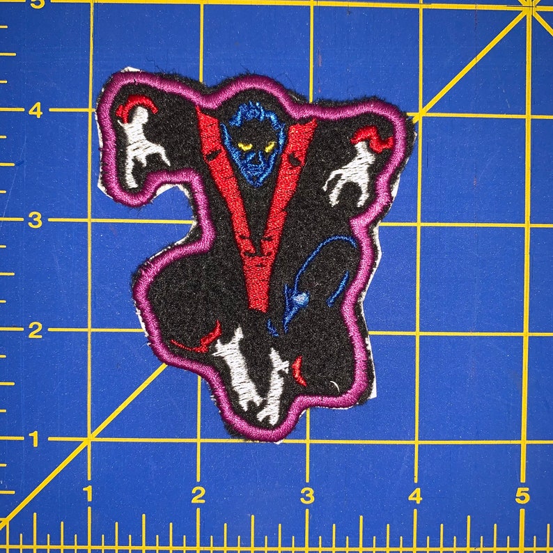 May include: A purple embroidered patch with a black background featuring a blue and yellow creature with red and blue accents. The creature is surrounded by white figures.