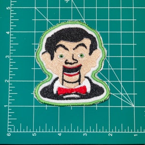 Puede incluir: Un parche bordado con un personaje de dibujos animados sonriente con cabello negro, una corbata de moño roja y un cuello blanco. El personaje tiene una gran sonrisa exagerada y ojos verdes. El parche está bordeado de verde y blanco.