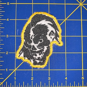 May include: Black and white embroidered patch of a grinning skull with a yellow outline. The skull has a large, toothy grin and one eye is visible.