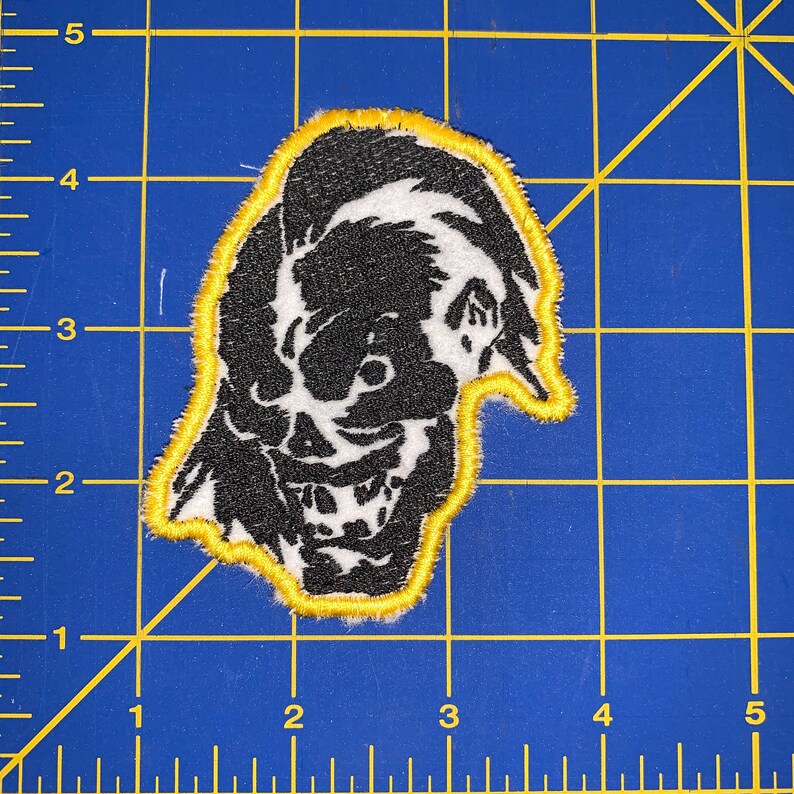 May include: Black and white embroidered patch of a grinning skull with a yellow outline. The skull has a large, toothy grin and one eye is visible.