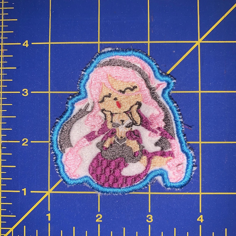 K&ouml;nnte beinhalten: Gestickter Aufn&auml;her mit einer Comicfigur mit rosa Haaren, die ein lila-schwarzes Outfit tr&auml;gt, mit einem blauen Umriss.