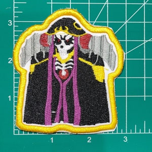 May include: Embroidered iron-on patch featuring a black and white character with a purple robe and yellow accents. The character has a skull-like face with red eyes and a crown.