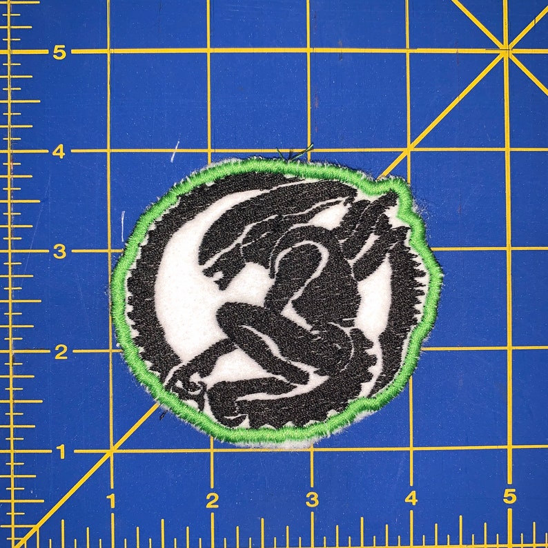 May include: An embroidered patch featuring a black and white alien creature, with a green border. The alien is crouched down with its arms raised above its head.