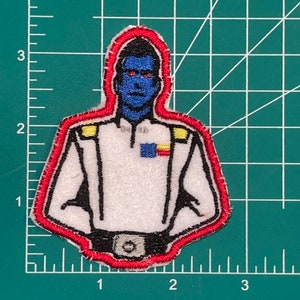May include: Embroidered patch of a blue-skinned person wearing a white uniform with red and yellow accents. The person has a serious expression and is wearing a black belt.