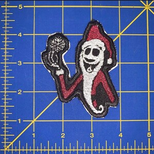 Puede incluir: Un parche de hierro bordado que presenta una figura esquelética blanca con un gorro de Papá Noel rojo y sosteniendo un copo de nieve. La figura tiene una amplia sonrisa y una larga barba blanca.