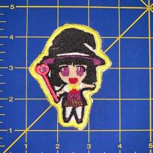 May include: Embroidered patch featuring a cartoon witch with black hair, a purple hat, and a pink wand. She is wearing a yellow, red, and purple outfit and white stockings. The patch has a yellow outline.