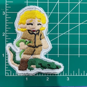 Pode incluir: Um remendo bordado com um personagem de desenho animado vestindo uma camisa marrom e segurando um jacaré verde. O personagem tem cabelo loiro e está sorrindo.