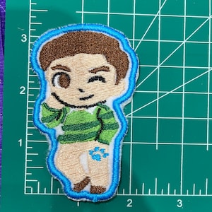 May include: Embroidered patch of a cartoon character with brown hair, a green and white striped shirt, and a blue outline. The character is winking and has a playful expression.