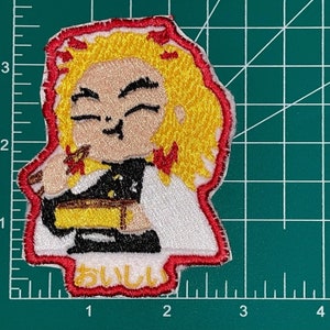 May include: Embroidered patch featuring a cartoon character with yellow hair, wearing a black and white outfit. The character is smiling and holding a wooden stick. The patch has a red border and the text "おいしい" in Japanese.