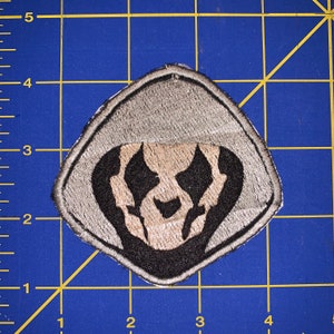 May include: A grey embroidered patch featuring a black hooded figure. The figure has a white face with black eyes and a black mouth.