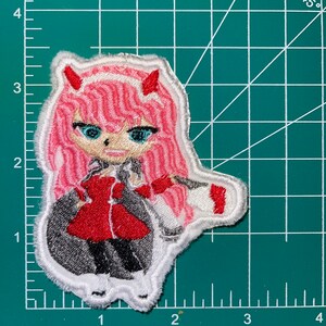 May include: Embroidered patch featuring a pink-haired anime character with red horns and a red jacket. The character is wearing a white collar and a white skirt. The patch is surrounded by a white border.