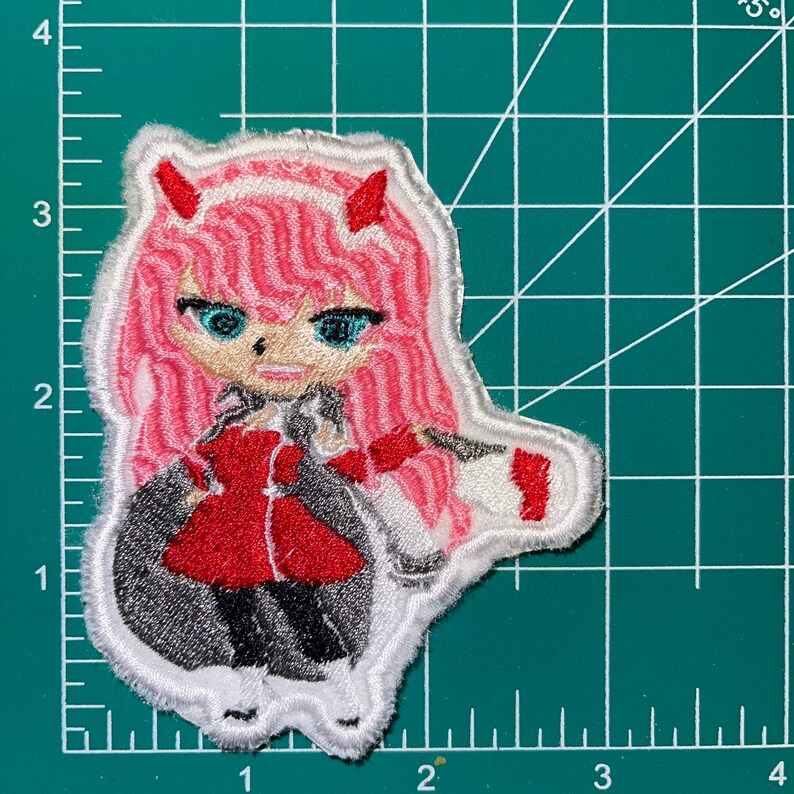 May include: Embroidered patch featuring a pink-haired anime character with red horns and a red jacket. The character is wearing a white collar and a white skirt. The patch is surrounded by a white border.